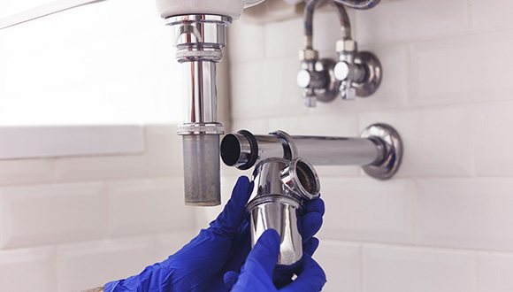 person wearing blue gloves inspecting plumbing under a sink with plumbing parts in focus and showing maintenance for efficient drainage system